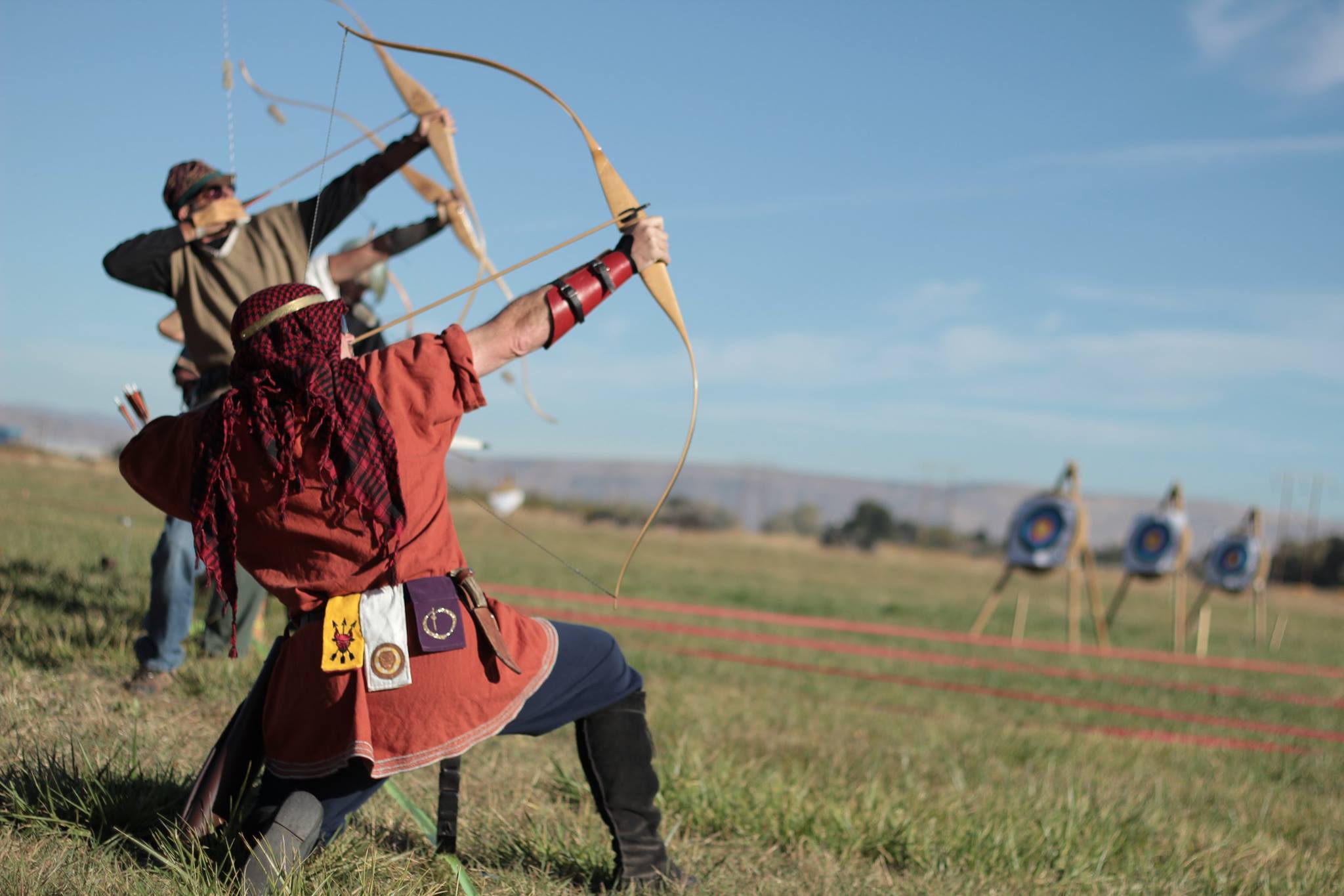 Image of a line of archers, in the foreground an archer in a red tunic and red and black head cover draws an arrow.